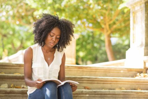 Focused beautiful young black woman reading book in park. Girl sitting on stairs with blurred green view in background. Park relaxation and reading concept. Front view.