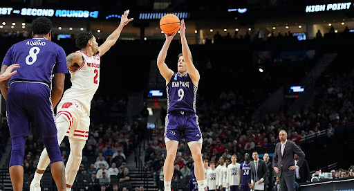 A man in a purple and white basketball jersey takes a shot while a man in a white and red jersey tries to block the shot during a High Point University men's basketball game.
