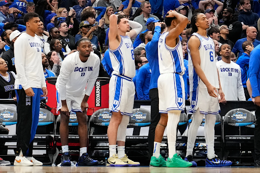 Several young men in white and blue basketball jerseys from the Duke University men's team stand on the sidelines of a basketball court watching the game.